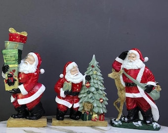 May include: Three Santa Claus figurines, each with a different pose and accessories. One Santa is carrying a stack of presents, another is holding a Christmas tree, and the third is carrying a bag and a reindeer. The figurines are made of painted resin and have a festive Christmas theme.