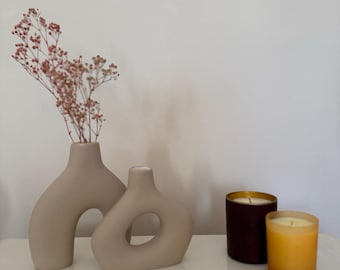 May include: Two beige ceramic vases with abstract shapes, one with a dried flower arrangement, and two candles, one burgundy and one yellow, on a white surface.