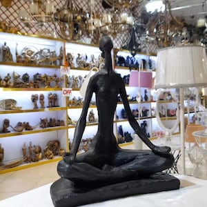 May include: Black, abstract sculpture of a person sitting in a meditative pose. The sculpture is made of a smooth, dark material and has a simple, elegant design.