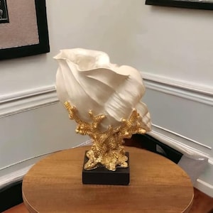 May include: A decorative seashell sculpture with a cream-colored shell and gold-colored coral-like base. The sculpture is mounted on a black rectangular base and sits on a wooden table.