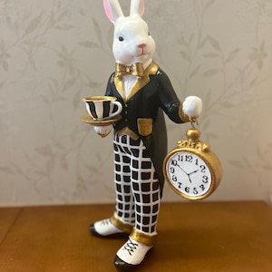 Può includere: Statuetta di coniglio bianco vestito con uno smoking nero con finiture dorate, che tiene in mano una tazza da tè e un orologio da tasca. Il coniglio indossa un papillon dorato e pantaloni a scacchi. L'orologio da tasca ha una cornice dorata e un quadrante bianco.
