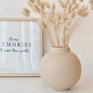 Könnte beinhalten: Eine beige Keramikvase mit rundem Korpus und einer breiten Öffnung, gefüllt mit getrocknetem Pampasgras. Die Vase steht auf einer weißen Oberfläche vor einem gerahmten Druck, auf dem "Make Memories all over the world." steht.