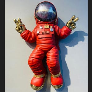 May include: A red astronaut figurine with a gold helmet and peace signs. The astronaut is wearing a red spacesuit with gold accents.