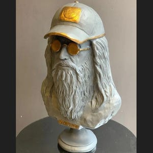 May include: A gray bust of a man with long hair and a beard, wearing a gray baseball cap with a gold emblem and gold-rimmed glasses. The bust is inscribed with "DA VINCI" at the base.