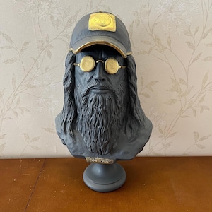 Könnte beinhalten: Eine dunkelgraue Büste von Leonardo da Vinci mit langem Haar und Vollbart. Die Figur trägt eine dunkelgraue Baseballkappe mit einem goldfarbenen Aufnäher und eine goldfarbene runde Brille. Der Sockel ist mit dem Text DA VINCI in Gold versehen.