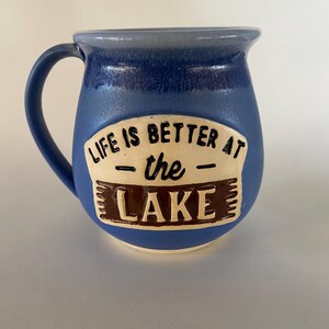 May include: A blue ceramic mug with a white rim and a brown and white sign that reads "Life is better at the lake".