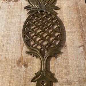 May include: A gold-toned pineapple-shaped trivet with a decorative, openwork design. The trivet has a loop on the bottom for hanging.
