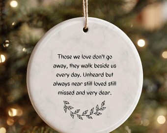 May include: A white ceramic Christmas ornament with a quote about loved ones. The text reads: "Those we love don't go away, they walk beside us every day. Unheard but always near still loved still missed and very dear."