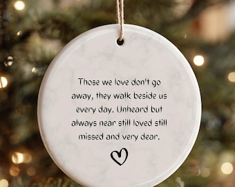 May include: A white ceramic ornament with a heart and the text "Those we love don't go away, they walk beside us every day. Unheard but always near still loved still missed and very dear." A brown string is attached.