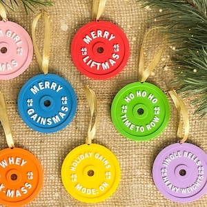 Könnte beinhalten: Eine Sammlung bunter Weihnachtsbaumschmuck-Gewichtsscheiben mit goldenen Bändern zum Aufhängen. Jedes Ornament zeigt einen anderen Fitness-Themen-Spruch, wie "Gym Girl X-Mas" und "Merry Liftmas", und das Gewicht "45 LB".