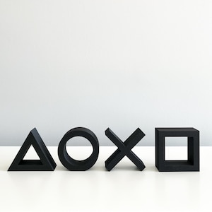 May include: Four black, 3D printed, geometric shapes: a triangle, a circle, an X, and a square. The shapes are arranged in a row on a white surface.