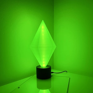 Op de afbeelding: Een groene diamantvormige lamp met een zwarte voet. De lamp is aan en geeft een groene gloed.
