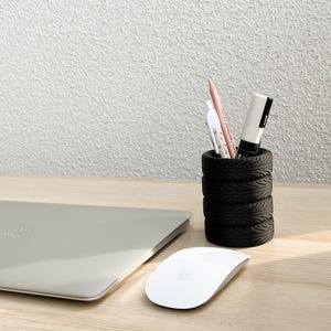 May include: A black tire-shaped pencil holder with a white marker, a pink pencil, and a white pen inside. The holder is on a light wood surface next to a silver laptop and a white wireless mouse.