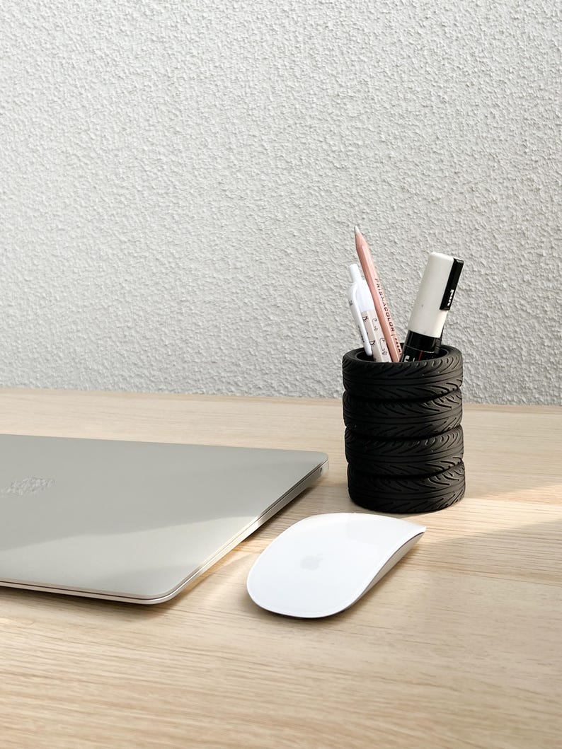 May include: A black tire-shaped pencil holder with a white marker, a pink pencil, and a white pen inside. The holder is on a light wood surface next to a silver laptop and a white wireless mouse.