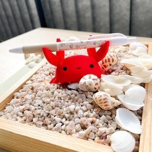 May include: A red crab-shaped pen holder with a white pen resting on top. The holder is surrounded by small white and brown seashells on a bed of white sand.