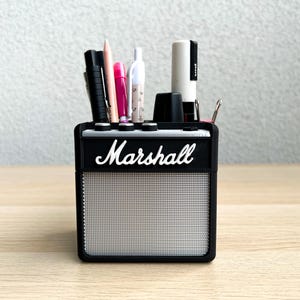 Könnte beinhalten: Schwarzer Schreibtisch-Organizer in Form eines kleinen Verstärkers mit einem silbernen Lautsprechergitter. Der Organizer ist mit dem Wort 'Marshall' gekennzeichnet und mit verschiedenen Schreibutensilien gefüllt, darunter Stifte, Bleistifte und Marker.