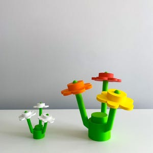 K&ouml;nnte beinhalten: Drei bunte Blumenskulpturen aus Kunststoff. Die Blumen sind rot, orange und gelb. Die Stiele sind gr&uuml;n und die Basis ist gr&uuml;n. Die Blumen sind in einer Gruppe angeordnet.