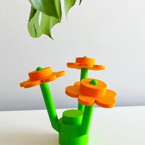 K&ouml;nnte beinhalten: Eine leuchtend gr&uuml;ne Keramikvase mit drei orangefarbenen, blumenf&ouml;rmigen Oberteilen. Die Vase hat einen abgerundeten Boden und drei Stiele, die sich nach oben erstrecken, um die Blumenoberteile zu st&uuml;tzen.