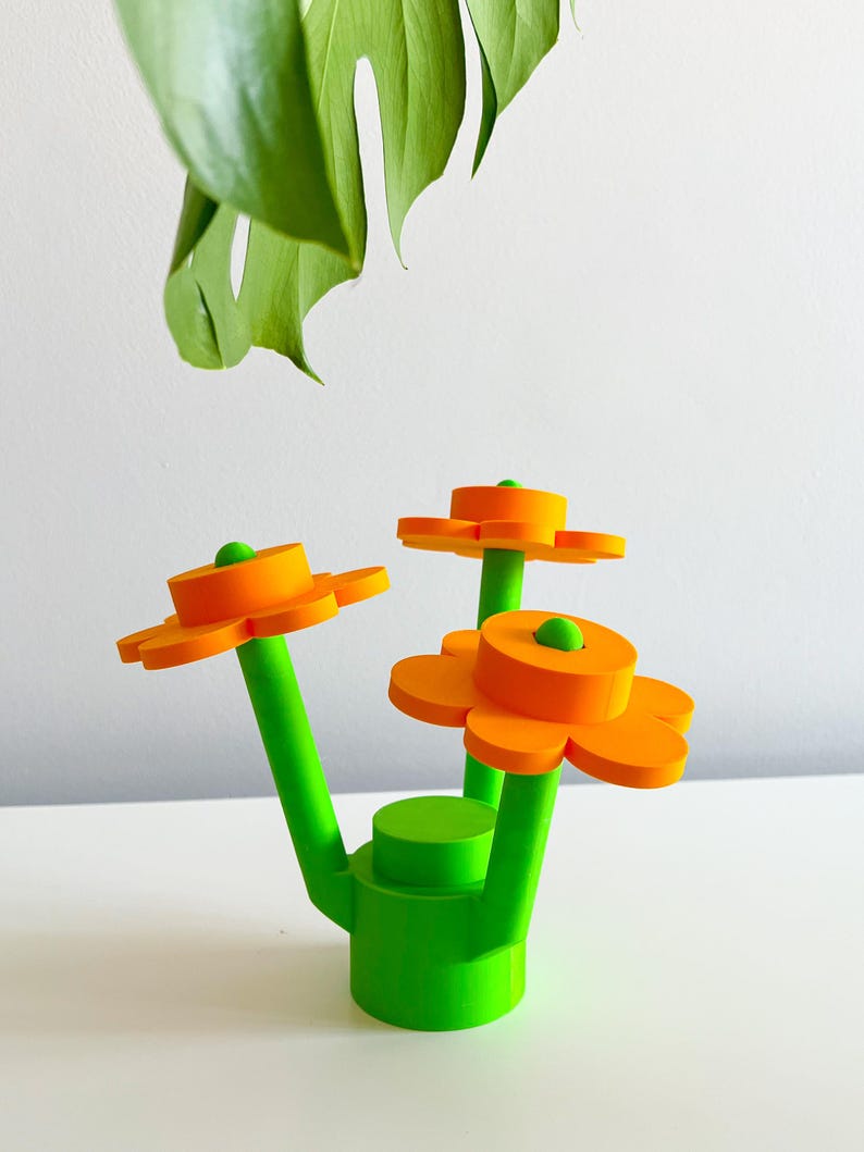 K&ouml;nnte beinhalten: Eine leuchtend gr&uuml;ne Keramikvase mit drei orangefarbenen, blumenf&ouml;rmigen Oberteilen. Die Vase hat einen abgerundeten Boden und drei Stiele, die sich nach oben erstrecken, um die Blumenoberteile zu st&uuml;tzen.
