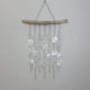 May include: A wall hanging composed of white, circular shell pieces suspended from a natural driftwood branch. The shell pieces are linked by silver-coloured chains, creating a cascading pattern. The driftwood is suspended from a silver chain, creating a decorative effect.