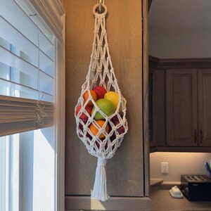 Peut inclure: Un porte-fruits en macramé blanc, suspendu à un anneau en bois, est rempli de fruits frais. Le design en coton tissé présente un pompon décoratif à la base. Les fruits comprennent des oranges, des pommes et des citrons.