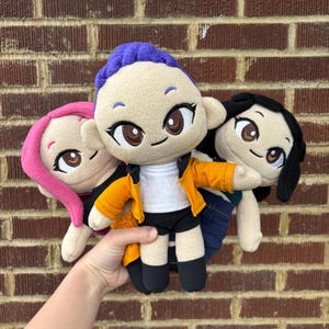 May include: Three plush dolls with large eyes and various hairstyles are held in front of a brick wall. The dolls have different outfits, including a yellow jacket, black shorts, and blue jeans. One doll has purple hair, another pink, and the third black.
