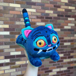 May include: A plush toy shaped like a cat with dark blue fur, bright yellow eyes, pink inner ears, and two white fangs. The creature features a striped tail with alternating dark blue and light blue bands and dark blue markings on its back.