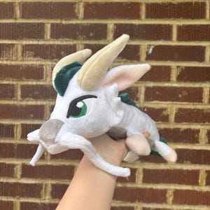 May include: A white plush dragon with cream-colored horns, green eyes, and green accents. The dragon has a long body and is being held up by a hand. The background is a brick wall.