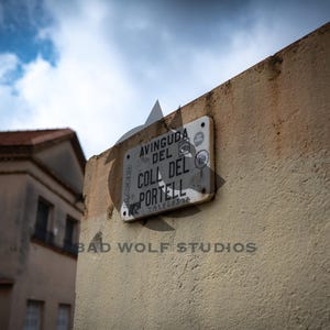 May include: A weathered street sign on a beige wall reads "AVINGUDA DEL COLL DEL PORTELL". The sign is rectangular, with faded text and some stickers. A building with a red tile roof is visible in the background.