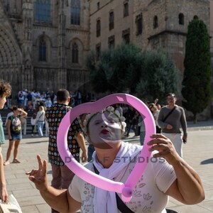 Puede incluir: Una persona con pintura facial sostiene un globo rosa en forma de corazón frente a su rostro. La persona lleva una camisa blanca y una corona de flores. Un gran edificio de piedra y una multitud de personas están en el fondo.