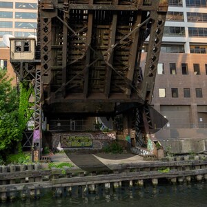 Può includere: Un ponte in metallo arrugginito sopra un canale con graffiti sui muri sottostanti. Il ponte è fatto di travi in acciaio e ha una passerella in legno. Il canale è circondato da muri di cemento e c'è vegetazione che cresce sulle rive.