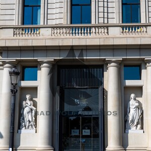 May include: Exterior view of a building with a classical architectural style. The facade features large windows with dark blue glass, a balcony, and decorative columns. Statues flank the entrance, and the building's name is visible on the glass doors.