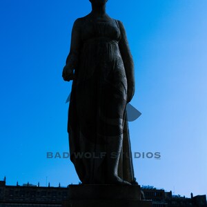 May include: A dark silhouette of a classical statue against a bright blue sky. The statue is of a woman in a flowing gown, standing on a stone pedestal. The text "BAD WOLF STUDIOS" is visible at the bottom.