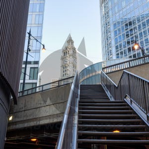 May include: A set of metal stairs leading up to a walkway with a view of a tall building with a clock tower in the background.