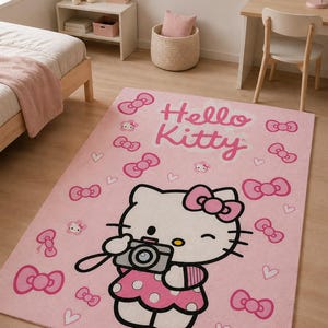 Può includere: Un tappeto rettangolare rosa con una grande grafica di Hello Kitty che tiene in mano una macchina fotografica, circondata da un motivo ripetuto di fiocchi rosa e piccoli cuori. Il testo Hello Kitty è stampato in corsivo in alto.