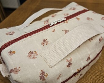 May include: A cream-colored, rectangular bag with a floral pattern in shades of pink and brown. The bag features a burgundy zipper and trim, a white zipper pull, and a white handle. The bag is made of quilted fabric.