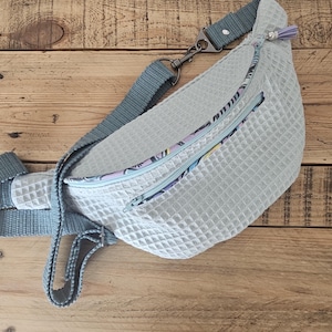 May include: A light gray fanny pack with a zippered pocket and a patterned fabric lining. The bag has a long adjustable strap.