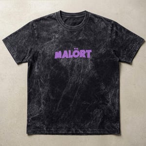 May include: Black t-shirt with a tie-dye effect and the word "MALÖRT" in purple, stylized font. The shirt has a crew neck and short sleeves. The fabric has a faded, vintage appearance.