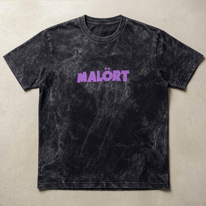 May include: Black t-shirt with a tie-dye effect and the word "MAL&Ouml;RT" in purple, stylized font. The shirt has a crew neck and short sleeves. The fabric has a faded, vintage look.