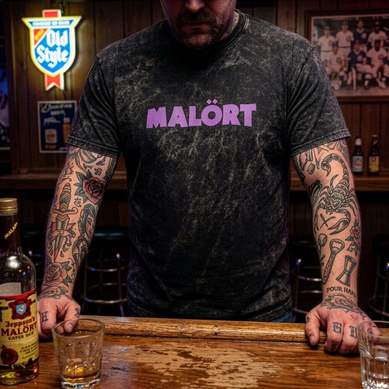 May include: A black t-shirt with the word "MAL&Ouml;RT" in purple block letters. The shirt has a textured appearance. A bottle of Jeppson's Mal&ouml;rt is on the bar. The person has tattoos on their arms.