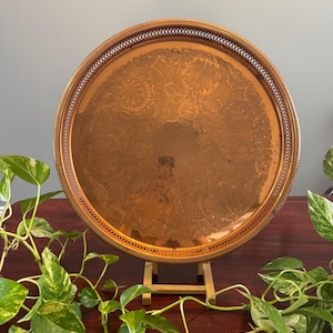 May include: A round copper serving tray with an ornate embossed design. The tray is on a wooden stand.