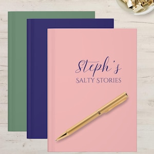 Puede incluir: Tres diarios de tapa dura en verde, azul marino y rosa. El diario rosa tiene el texto "Steph's SALTY STORIES" en una fuente cursiva, con un bolígrafo dorado encima. Un pequeño cuenco con clips dorados está en la esquina superior derecha.