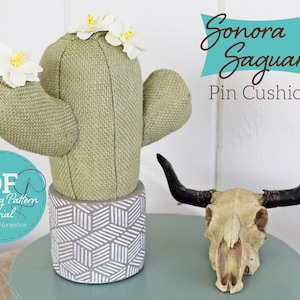 May include: A green burlap pincushion shaped like a saguaro cactus with white fabric flowers on top. The cactus is sitting on a white and gray geometric patterned pot. A white cow skull with black horns is sitting on a light blue surface. The text "Sonora Saguaro Pin Cushion" is written in brown text on a teal blue background.