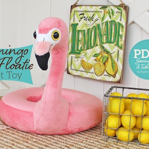 Puede incluir: Juguete de peluche rosa en forma de flotador de flamenco con pico negro y ojos amarillos. El juguete está sentado en una alfombra tejida frente a una pared blanca. Un letrero que dice "Fresh Lemonade" está colgado en la pared. Una cesta de alambre llena de limones está en el lado derecho de la imagen. El texto "PDF Sewing Pattern & Tutorial by Rustic Hoosier" está en un círculo azul en el lado derecho de la imagen.