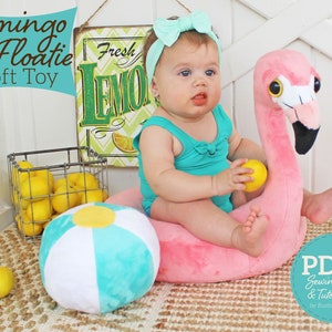 Puede incluir: Un bebé está sentado en un flotador de flamenco rosa, sosteniendo una pelota amarilla. La imagen incluye una pelota de playa, limones en una cesta de alambre y un letrero que dice "Fresh Lemon". El texto en la imagen dice "Flamingo Float Soft Toy".