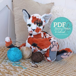 Op de afbeelding: Een PDF-naai-patroon en een tutorial voor het maken van een calico kattenknuffel. Het patroon is van Rustic Horseshoe. De afbeelding toont een afgewerkt calico kattenknuffel dat op een geweven tapijt zit met garenbollen.