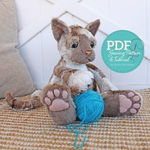 Puede incluir: Un juguete de peluche de gato con pelaje marrón y blanco, sentado con una bola de lana azul. El gato tiene una nariz rosa y almohadillas de patas rosas. La imagen incluye el texto "PDF Sewing Pattern & Tutorial by Rustic Horseshoe".