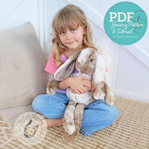 Puede incluir: Una niña pequeña está sentada en una alfombra tejida y sostiene un conejito de peluche marrón y blanco. El conejito tiene orejas largas y caídas y una textura suave y mullida. La niña lleva una camisa rosa y pantalones vaqueros azules. La imagen es un primer plano de la niña y el conejito.