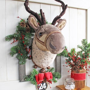 K&ouml;nnte beinhalten: Ein Pl&uuml;sch-Rentierkopf, der an einer Wand mit einem Weihnachtskranz und einer roten Schleife mit der Nummer 24 befestigt ist.