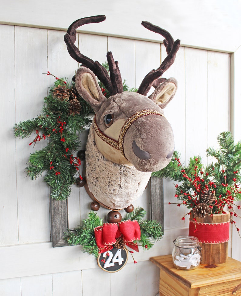 K&ouml;nnte beinhalten: Ein Pl&uuml;sch-Rentierkopf, der an einer Wand mit einem Weihnachtskranz und einer roten Schleife mit der Nummer 24 befestigt ist.